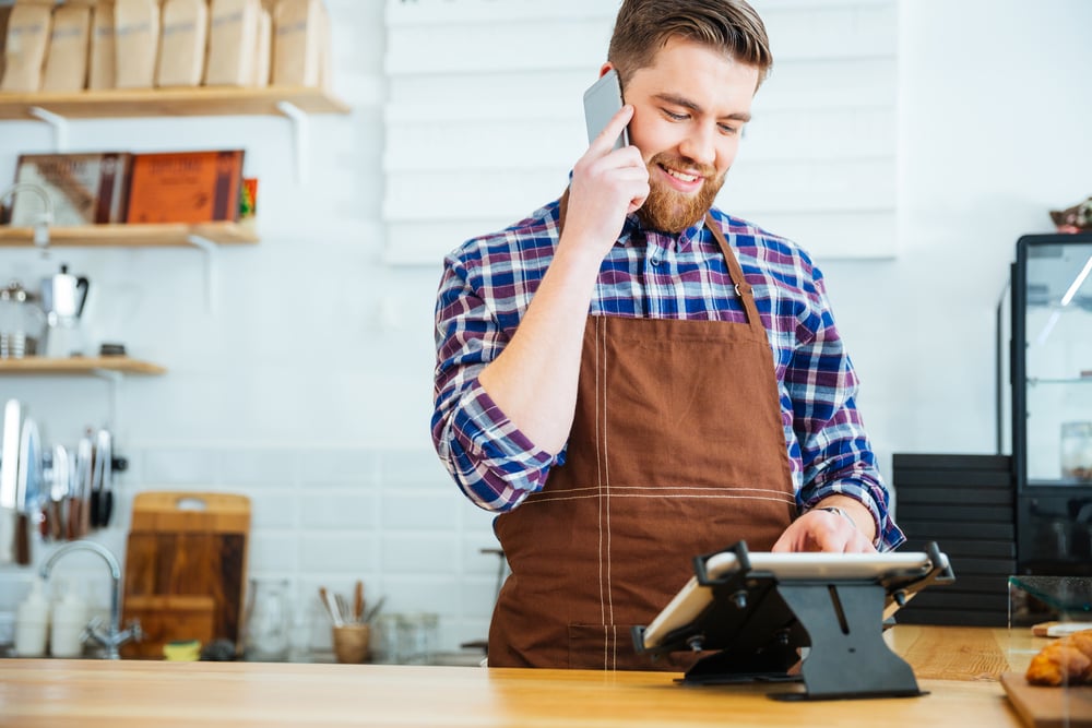 Handsome smiling barista with beard taking order on cell phone and using tablet in cafeteria Handsome smiling barista with beard taking order on cell phone and using tablet in cafeteria